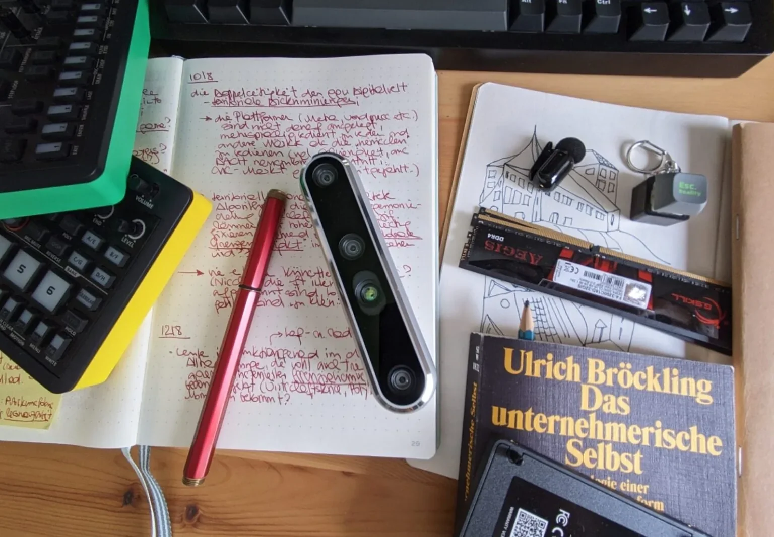 A flatlay photograph of various items relating to artistic research and digital culture: notebooks with scribbled notes and drawings, a copy of Ulrich Bröckling's monograph "Das unternehmerische Selbst" with a pen stuck inside, a fountain pen, two colourful Roland Aira synthesizers, a keyboard, a webcam, two RAM sticks and a clip-on microphone.